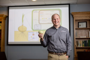A man in a gray shirt holds an onion and stands in front of a projection showing a diagram of an onion's biological processes during Investors Day in an Innovation District library or office setting.