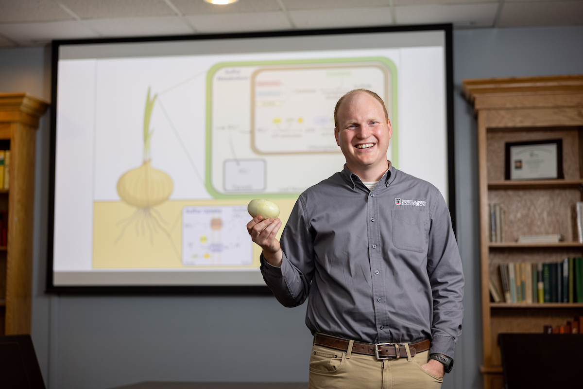 A man in a gray shirt holds an onion and stands in front of a projection showing a diagram of an onion's biological processes during Investors Day in an Innovation District library or office setting.