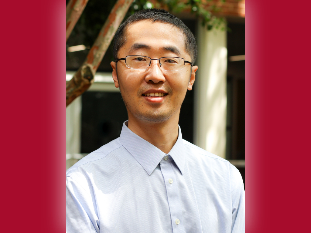 A man with glasses and short hair, wearing a light blue button-up shirt, is smiling outdoors in front of a building with a blurred background—Li, recipient of the Young Investigator Award.