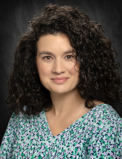 Lauren Lazaro wears a blue and green patterned blouse while posing in front of a dark, blurred background.