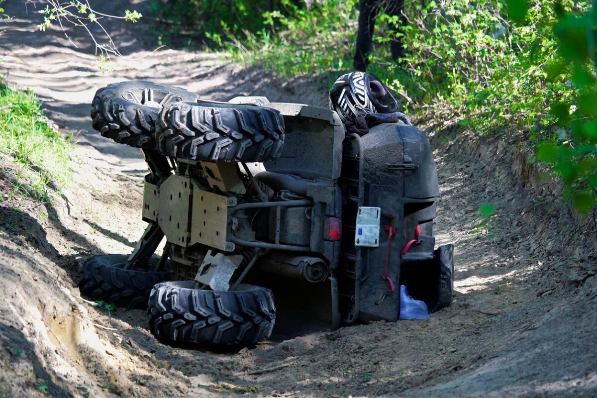 An overturned ATV lies on its side in a dirt path surrounded by greenery, with a helmet resting on the vehicle—a vivid reminder of the importance of ATV safety.