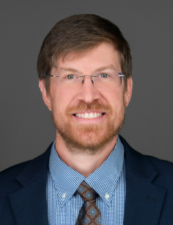 Brad Edenfield Man with light brown hair, beard, and glasses, wearing a blue suit jacket, patterned tie, and checkered shirt, smiling in front of a gray background.