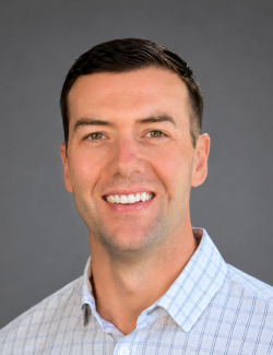 Bryce Wilberding A man with short dark hair, wearing a light plaid button-up shirt, smiles at the camera in front of a plain gray background.