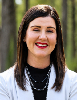Caitlin Jackson A woman with straight dark hair, wearing a light blazer, black top, and layered necklaces, smiles outdoors with blurred trees in the background.
