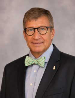 Donald Chase A man in a suit, light blue shirt, and green bow tie wearing glasses, poses in front of a plain gray background.