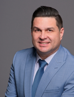 Juan F. DeVillena Man in a light blue suit jacket, white shirt, and blue tie, smiling at the camera against a plain gray background.