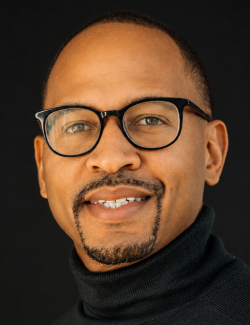 Michael Bailey A man wearing glasses and a black turtleneck smiles in front of a dark background.