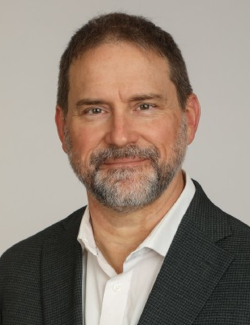 Mark Breutzman Middle-aged man with short brown hair and a gray beard, wearing a white shirt and dark blazer, posed against a plain background.