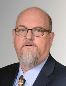 Michael Howell A middle-aged man with glasses, a short gray beard, and a receding hairline wearing a suit, dress shirt, and patterned tie against a neutral background.