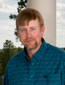Mark Rodgers A man with light brown hair and a goatee, wearing a blue plaid shirt, stands outdoors in front of a column and trees.