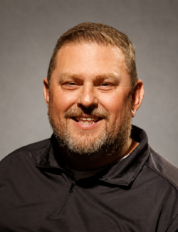 Matt Rushing Smiling middle-aged man with short hair and a beard, wearing a dark zip-up jacket, posing in front of a plain gray background.