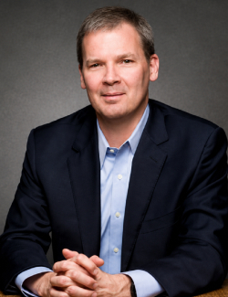 Mark Theuerkauf A man in a dark suit jacket and light blue shirt sits with hands clasped, facing the camera against a plain, dark background.