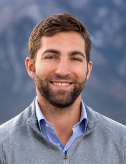 Nick Schroer A man with brown hair and a beard is smiling, wearing a light blue collared shirt and gray sweater, with a blurred outdoor background.