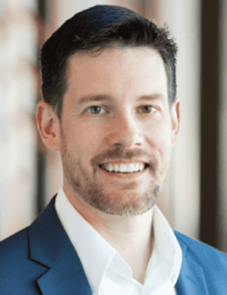Travis Frey A man with short dark hair and a beard, wearing a blue suit jacket and white shirt, smiling at the camera in an indoor setting.
