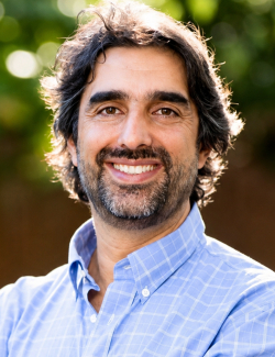A man with medium-length dark hair and a beard, wearing a blue checkered shirt, smiles at the camera outdoors with greenery in the background.
