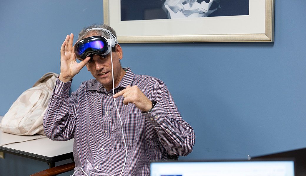Dr. Kyle Johnsen, wearing a checkered shirt, adjusts a virtual reality headset on their head while seated at a table in an office setting, embodying the spirit of innovation recognized by the Georgia Life Sciences Innovation Award.