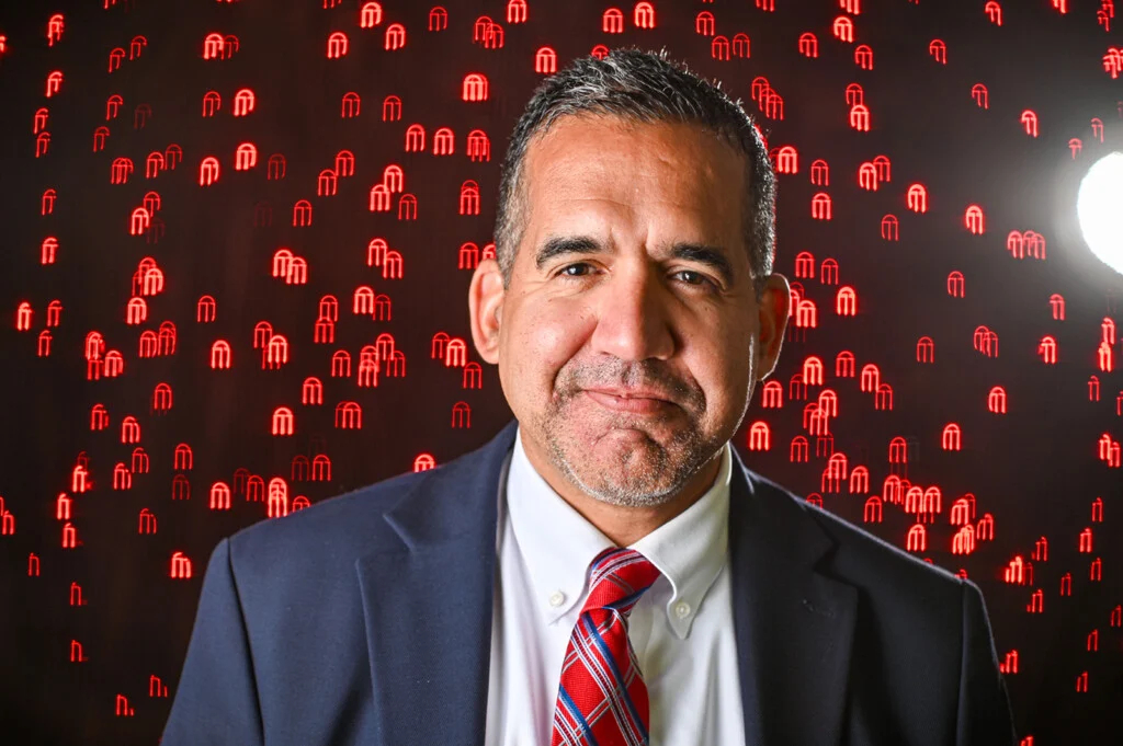 A man in a suit and red striped tie smiles slightly; red lights form M shapes in the blurred background, with a bright light source on the right, embodying the confidence often seen among UGA Leaders who drive research education forward.