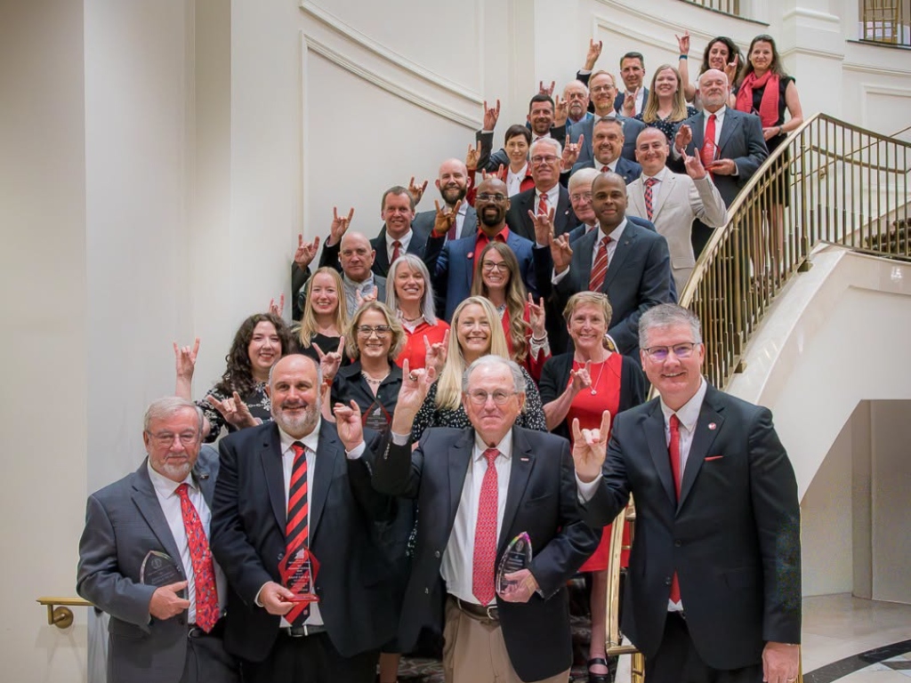 A large group of people in formal attire, including several NC State Alumni and IIPA Members, stand on a staircase—many holding awards and making a hand gesture—posing for a group photo indoors.