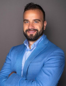 Man with short brown hair and beard wearing a light blue blazer and shirt, standing with arms crossed and smiling in front of a plain gray background. This is Konstantin Cvetanov.
